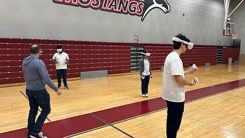 Three Nursing students in the Health Sciences gymnasium wearing white virtual reality headsets are practicing treating a patient. A staff member in a blue hooded sweatshirt oversees them.