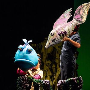 (From left) Kelly Grago, Mukta Phatak in a performance of the TheaterWorksUSA musical production of “The Pout-Pout Fish.” The actors are holding brightly colored fish puppets on stage.