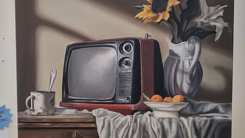 A still life oil painting of a TV with a yellow flower in a vase. They are placed on a white sheet on top of a wooden table. There's a mug with a spoon sticking out of it. There's also a bowl of fruit near the vase.