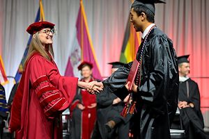 Dr. Vicki Bastecki-Perez dressed in red regalia shakes hands with graduates on stage at Commencement