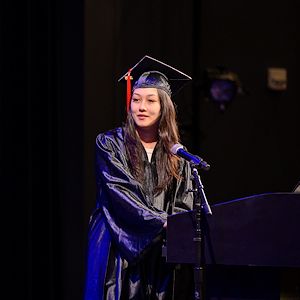 Gateway to College graduate Cat Cave dressed in a black cap and gown speaks at a lectern during the Gateway to College graduation on stage