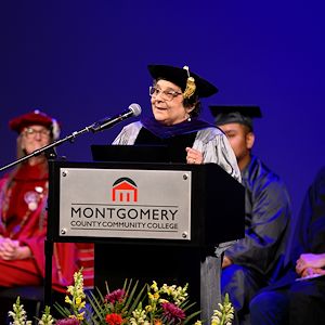 Dr. Rosann Bar, dressed in regalia, speaks at a lectern during the Gateway to College graduation ceremony on stage