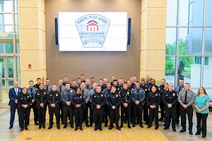 Municipal Police Academy Class 2601 stands in the Health Sciences Center atrium as a group. The TV screen above them says Municipal Police Academy