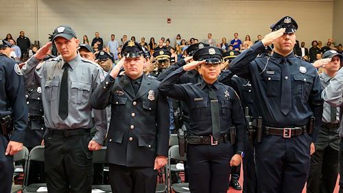 Graduating Municipal Police Academy cadets stand saluting at attention during the program's graduation ceremony inside the Educational Gym of the Health Sciences Center. A crowd of people sits behind them. 