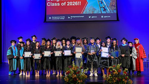 Students dressed in black graduation attire hold certificates on stage. Faculty in academic regalia stand next to them