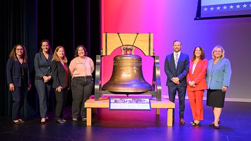 Seven people stand next on a stage next to a replica of the Justice Bell. 