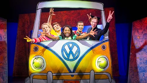 Five “Pete the Cat” actors on stage stand behind a cutout of a VW bug. They are flashing piece signs. 