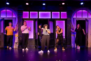 The Second City cast members perform a scene with several actors holding microphones on stage. 