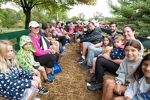 Children and adults sit in a hay-filled wagon during a hayride. Tall trees surround them. 