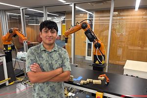 Heber Herrera standing in front of a robotic mechanical arm. His arms are crossed.