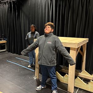 An actor rehearses a scene a scene of a play. His arms are spread widely, and his head is tilted up toward the ceiling. An actress stands behind him.