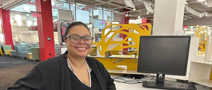 Kimberly Madera sitting at a computer in the library. She's seated wearing glasses and smiling.