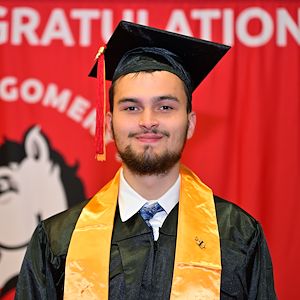 Aaron Laws in a cap and gown stands in front of a red Montgomery County Community College backdrop before Commencement