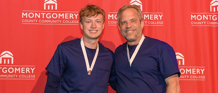 Father and son David and Aiden Gawel stand together wearing nursing scrubs in front of a red Montgomery County Community College backdrop