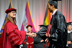 Dr. Bastecki-Perez dressed in red academica ragalia shakes Vincent Nguyen's hand during commencment. He's dressed in a black cap and gown.